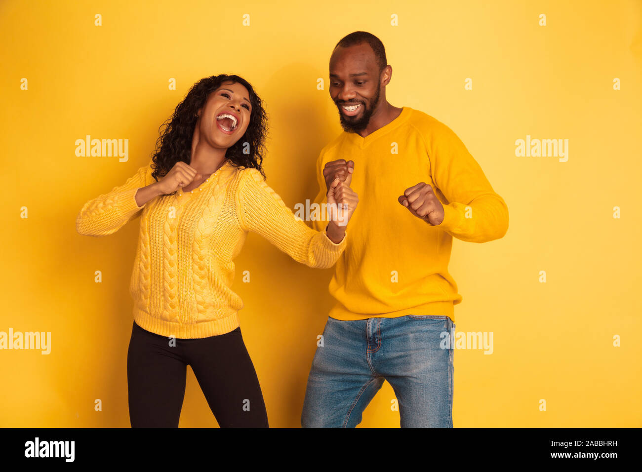 Young emotional african-american man and woman in bright casual clothes on yellow background. Beautiful couple. Concept of human emotions, facial expession, relations, ad. Dancing and singing. Stock Photo