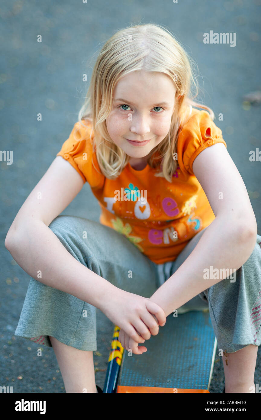 Casual, pretty blonde girl with enigmatic smile sitting on a skateboard and making eye contact Stock Photo