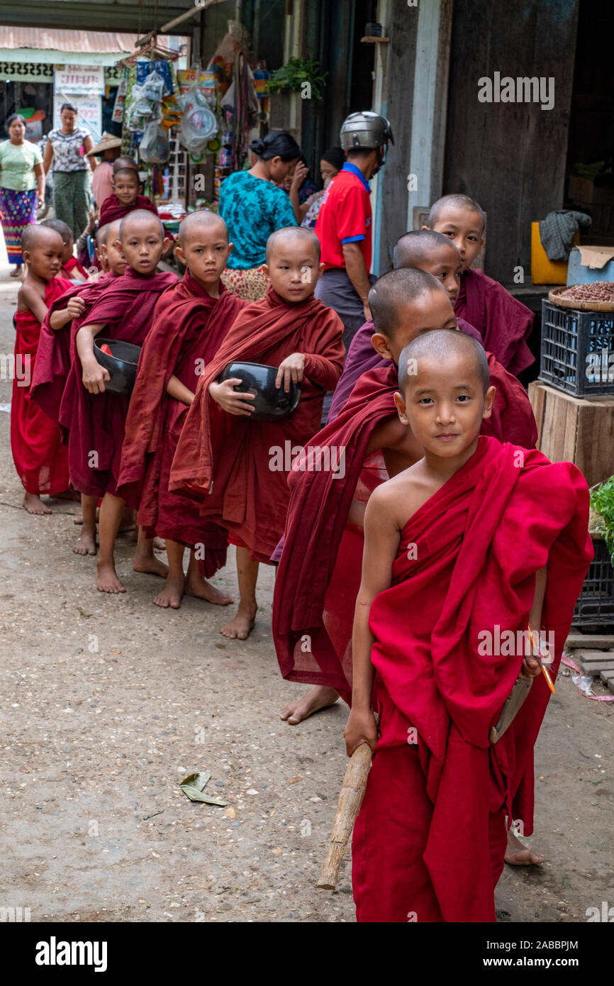Many young Buddhist monks dressed in scarlet robes enter the market of Kanne, Myanmar (Burma) in search of alms and led by a young monk with a gong Stock Photo