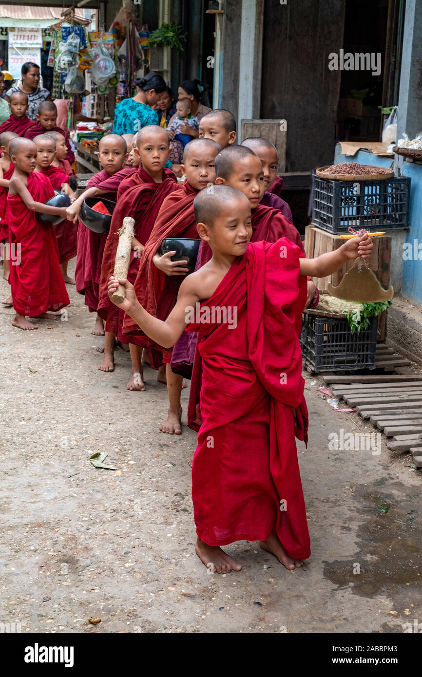 Many young Buddhist monks dressed in scarlet robes enter the market of Kanne, Myanmar (Burma) in search of alms and led by a young monk with a gong Stock Photo