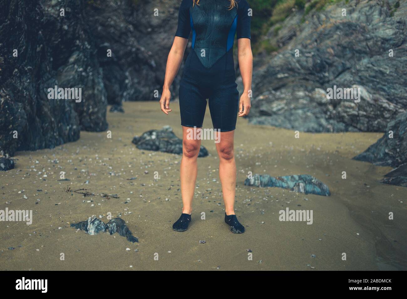 A young woman wearing a wetsuit is standing on a beach with some rocks