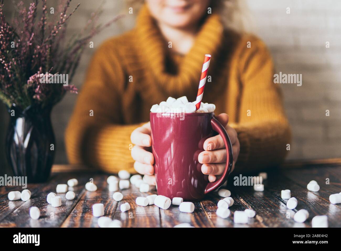 Woman in yellow sweater holding hot chocolate mug covered with marshmallow on dark wood background. Stock Photo