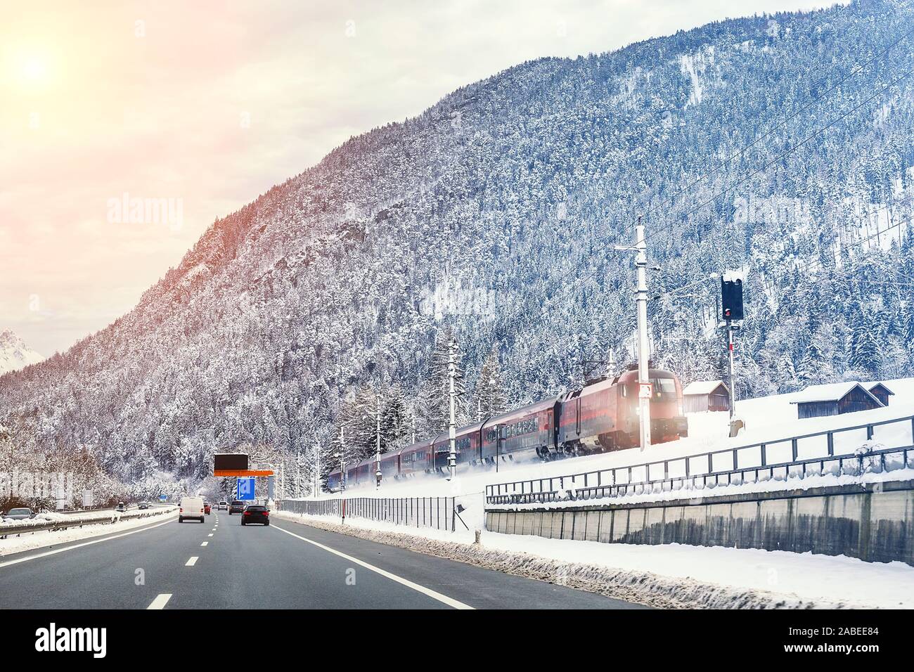 Winter alpine mountain highway and parallel railway road with intercity fast train and cloude sunset sky on background at cold day. Car trip family Stock Photo