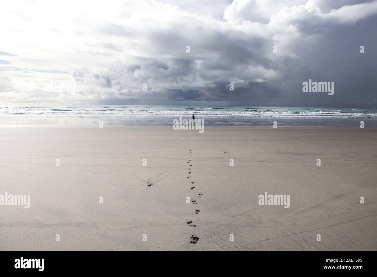 Woman in distance standing against rough sea on very long beach under dramatic cloudy sky with straight track of footprints leading to her. Fossil Poi Stock Photo