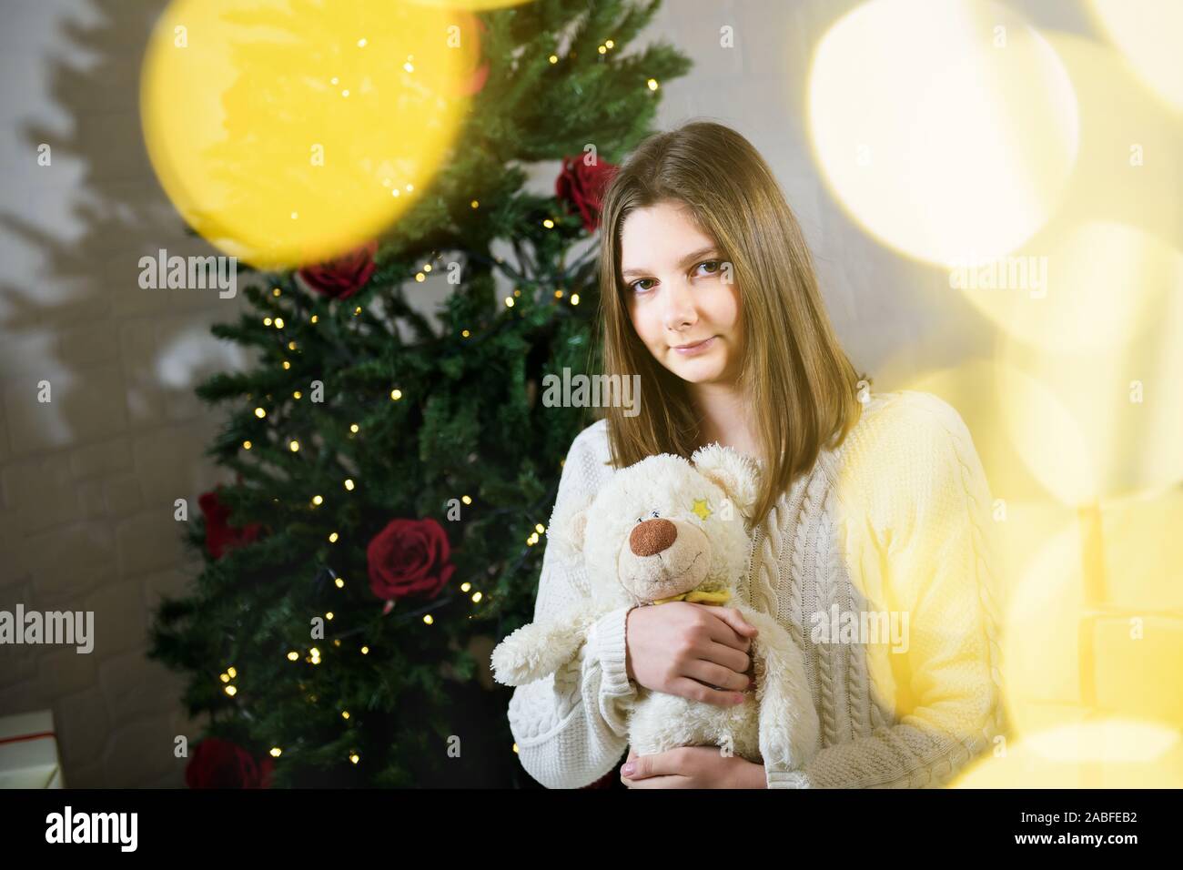 girl sits near a Christmas tree and holds in her hands a toy teddy bear Stock Photo