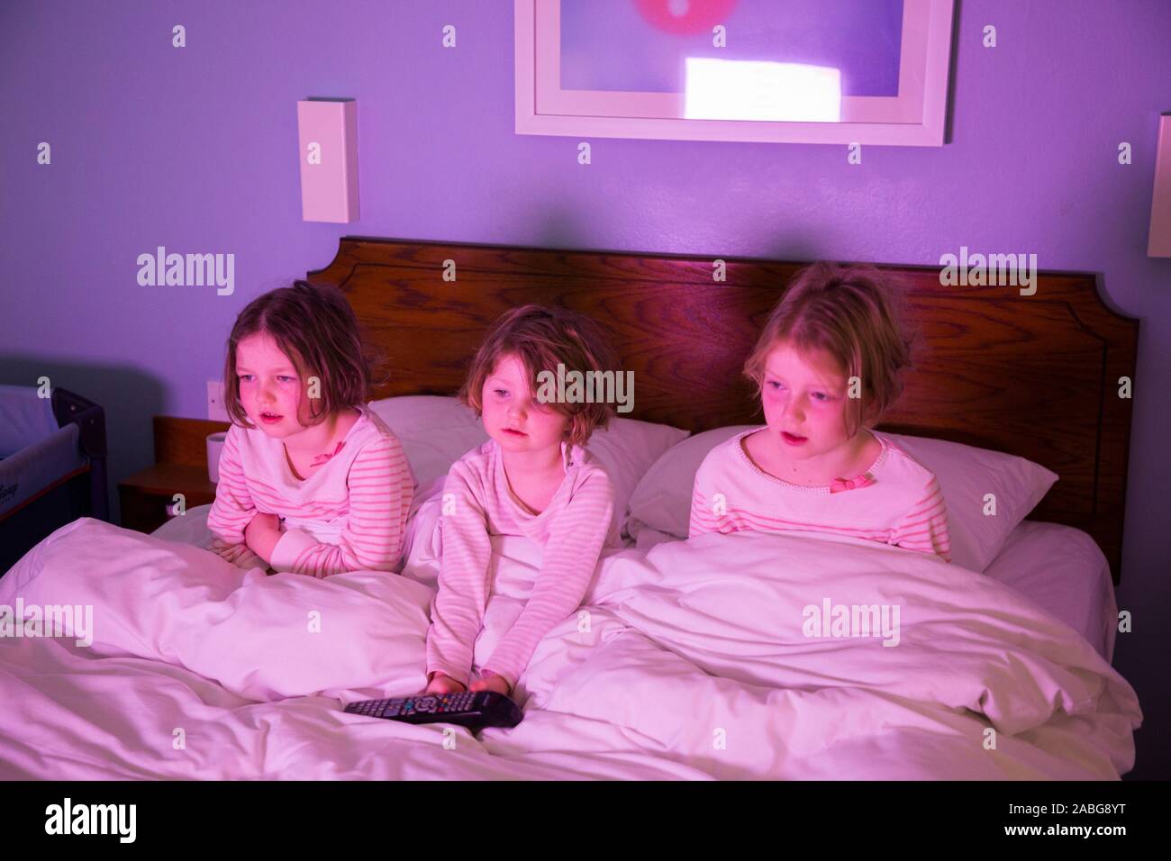 Three girls – aged 6, 4 and eight years old – in a hotel bed watching TV children's program on in the hotel television while on holiday, after waking up in the morning, but equally could be illustrating going to bed at night and watching TV.(105) Stock Photo