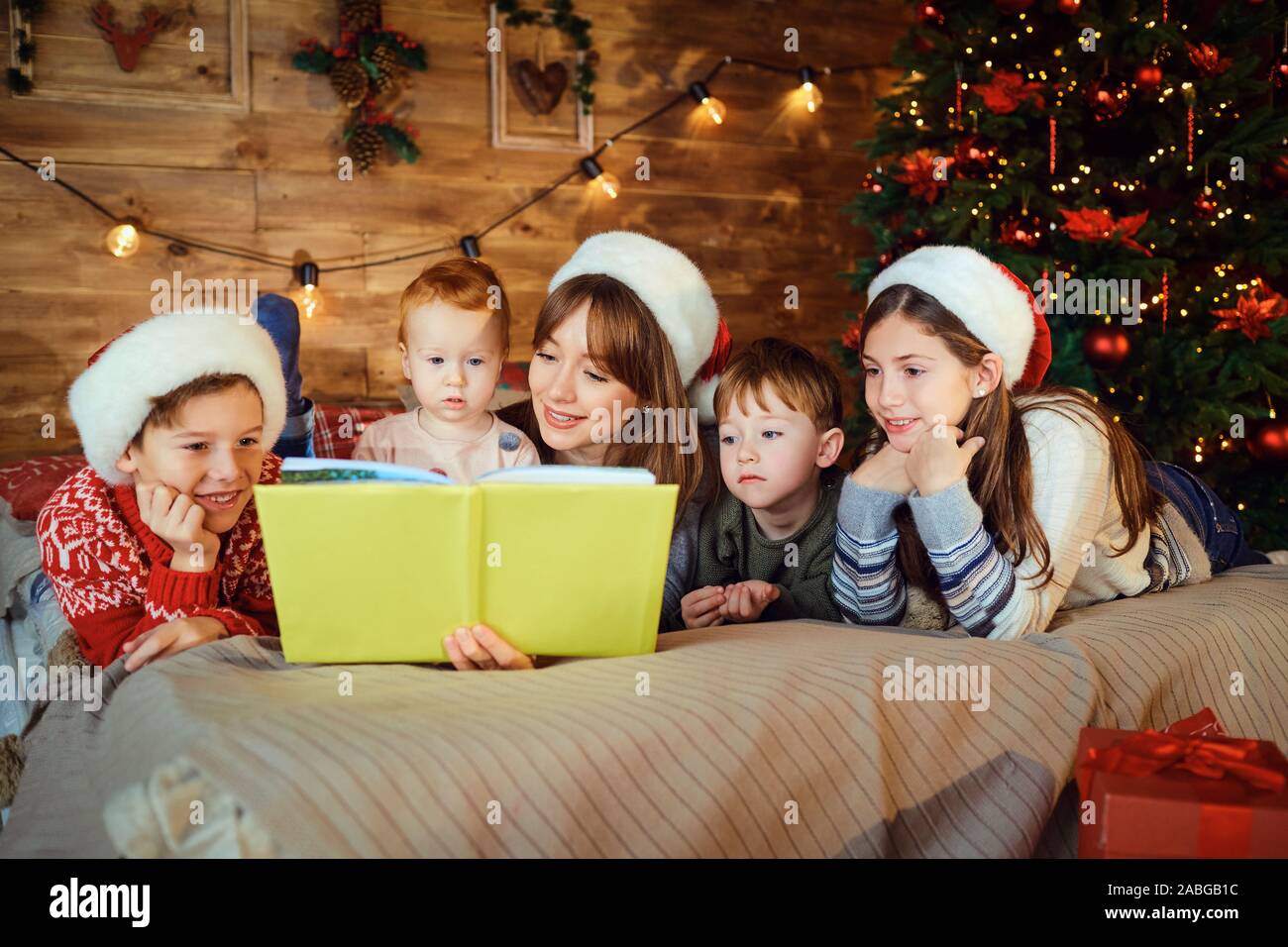 Mother reads a book with children lying on the bed in the room with tree at Christmas. Stock Photo