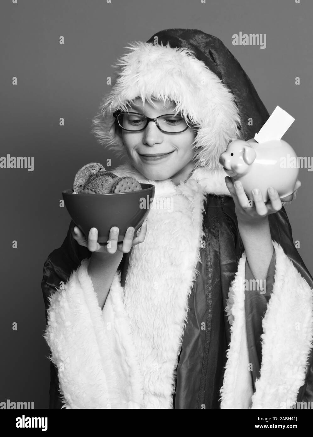 young cute santa claus boy with glasses in xmas sweater and new year christmas hat. holding pink piggy pig bank and chocolate hip cookies in bowl on red studio background. Stock Photo