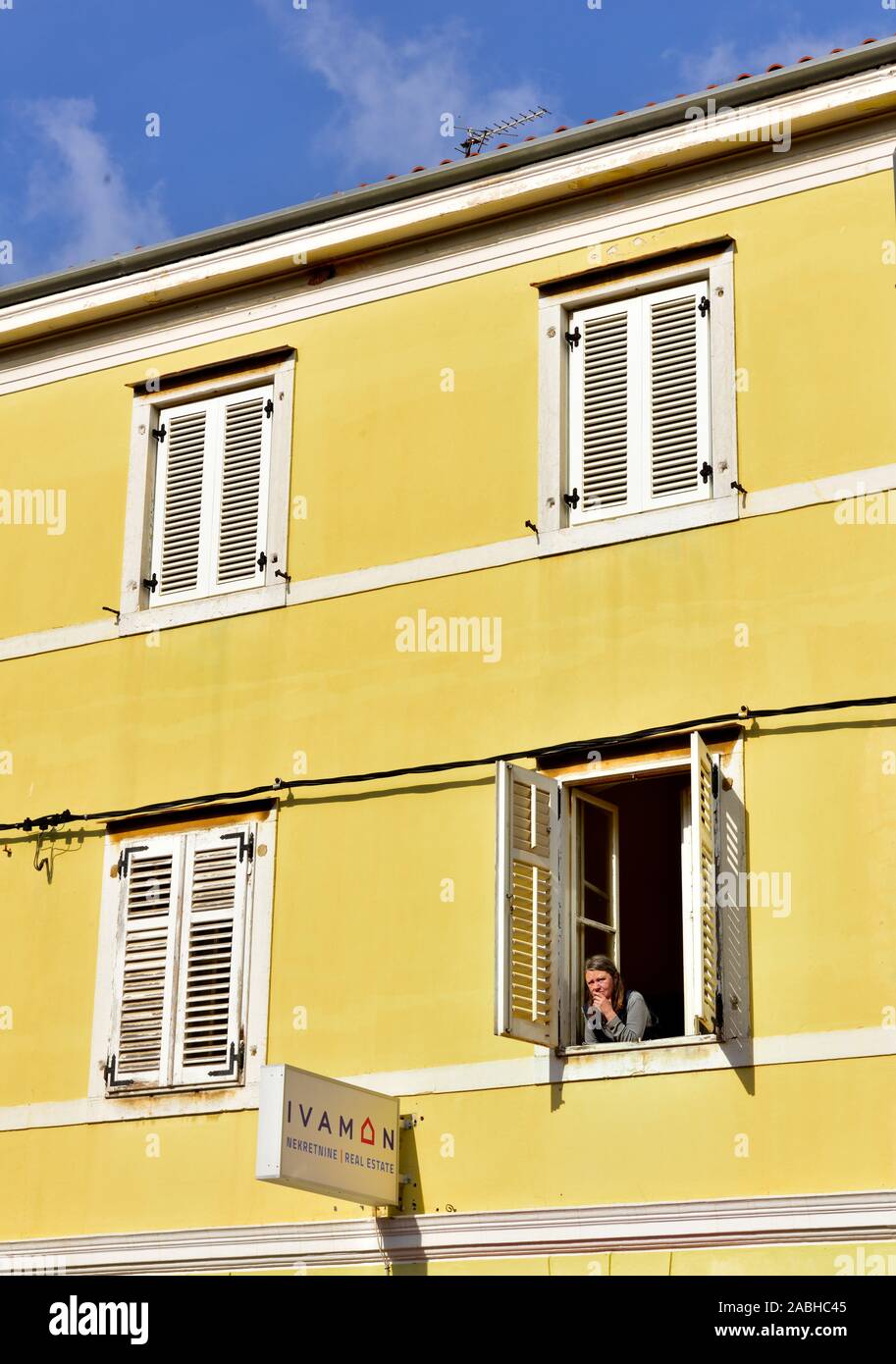 Looking up from outside at woman looking out of open window with shutters in yellow building Stock Photo