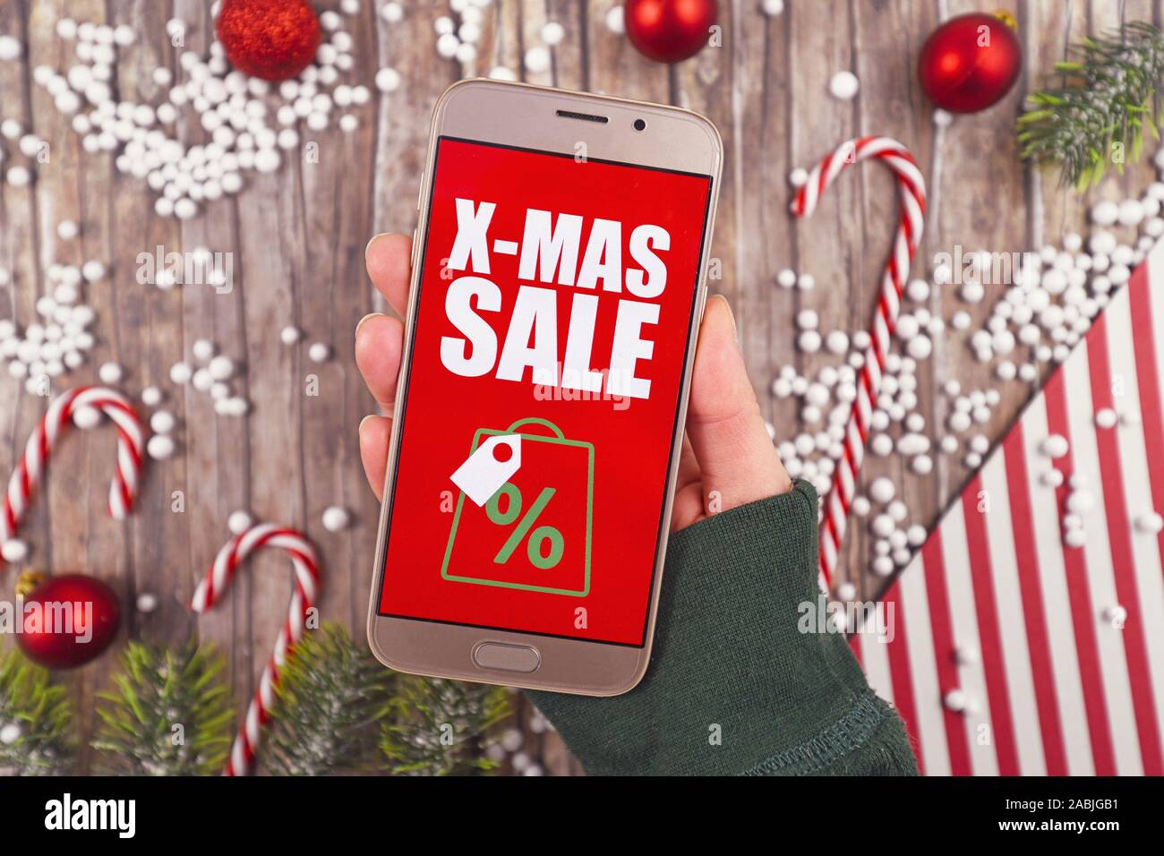Concept for Christmas seasonal online shopping and sales with hand holding cell phone with red 'X-Mas Sale' sign in front of desk with seasonal decora Stock Photo