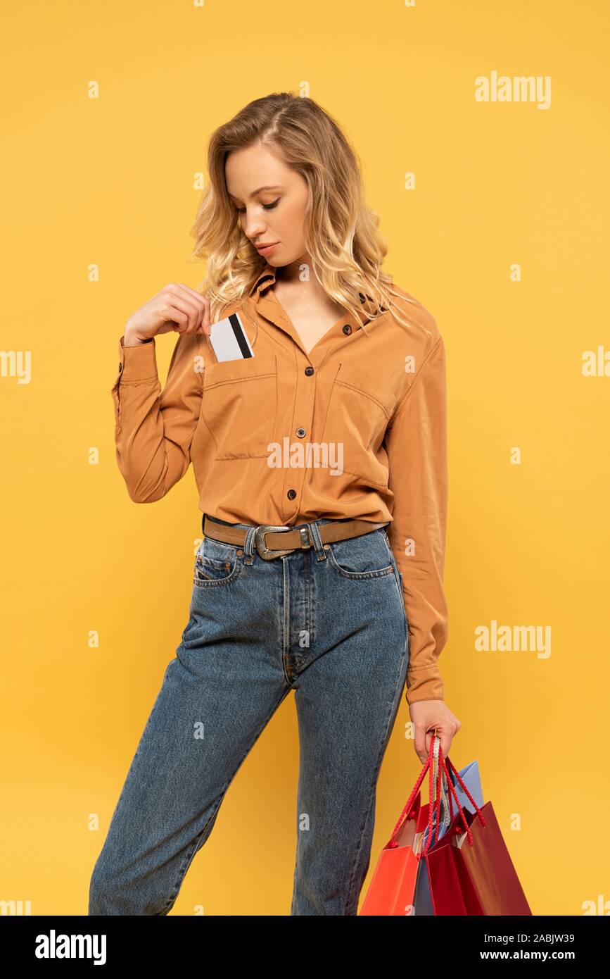 Blonde woman putting credit card in shirt pocket and holding shopping bags isolated on yellow Stock Photo