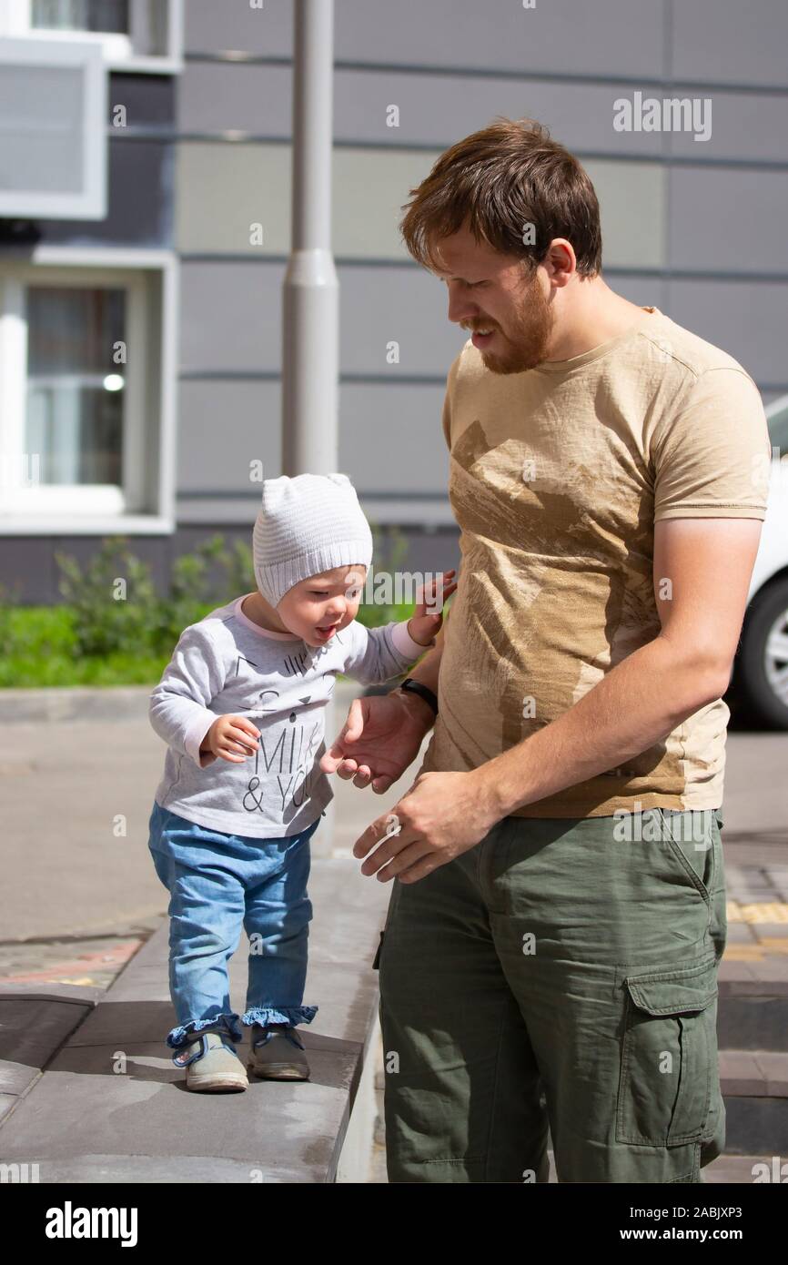A young father teaches his child to walk. Little girl a boy in jeans and a hat walks with dad. Family on a walk, warm sunny day in the city. Caucasian Stock Photo
