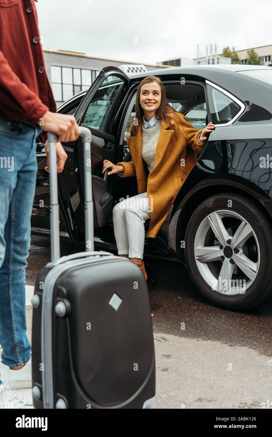 Driver holding wheeled suitcase and smiling woman sitting in taxi Stock Photo