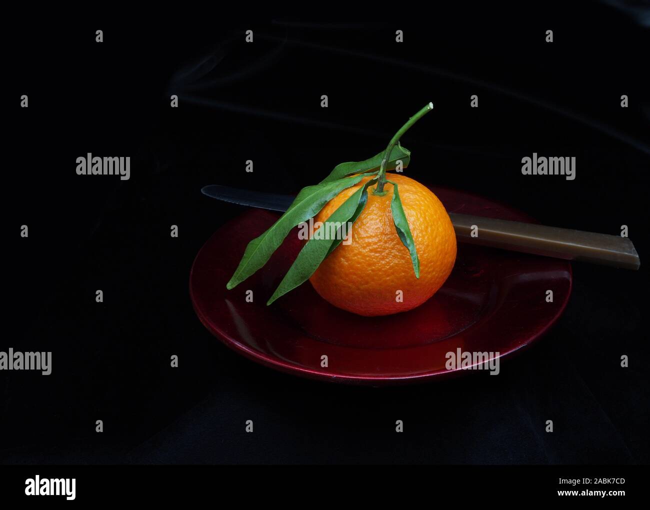 Mandarin type small orange on red plate, with leaves. Whole, unpeeled. With knife, dark background. Stock Photo