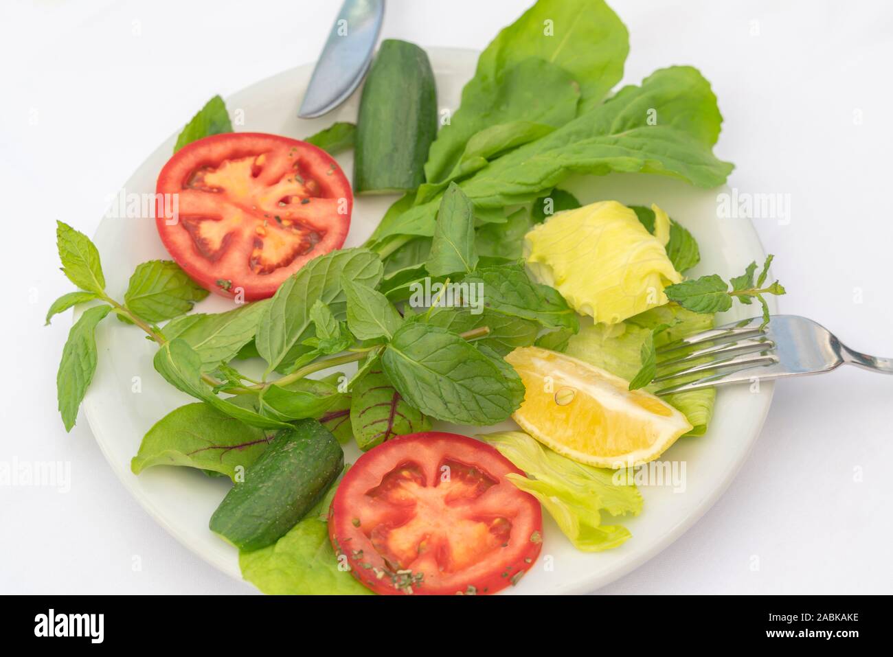 Salading from spinach, mint, mangold, greens, lemon, tomato and cucumber slices on the white plate with knife and fork on white background. Stock Photo