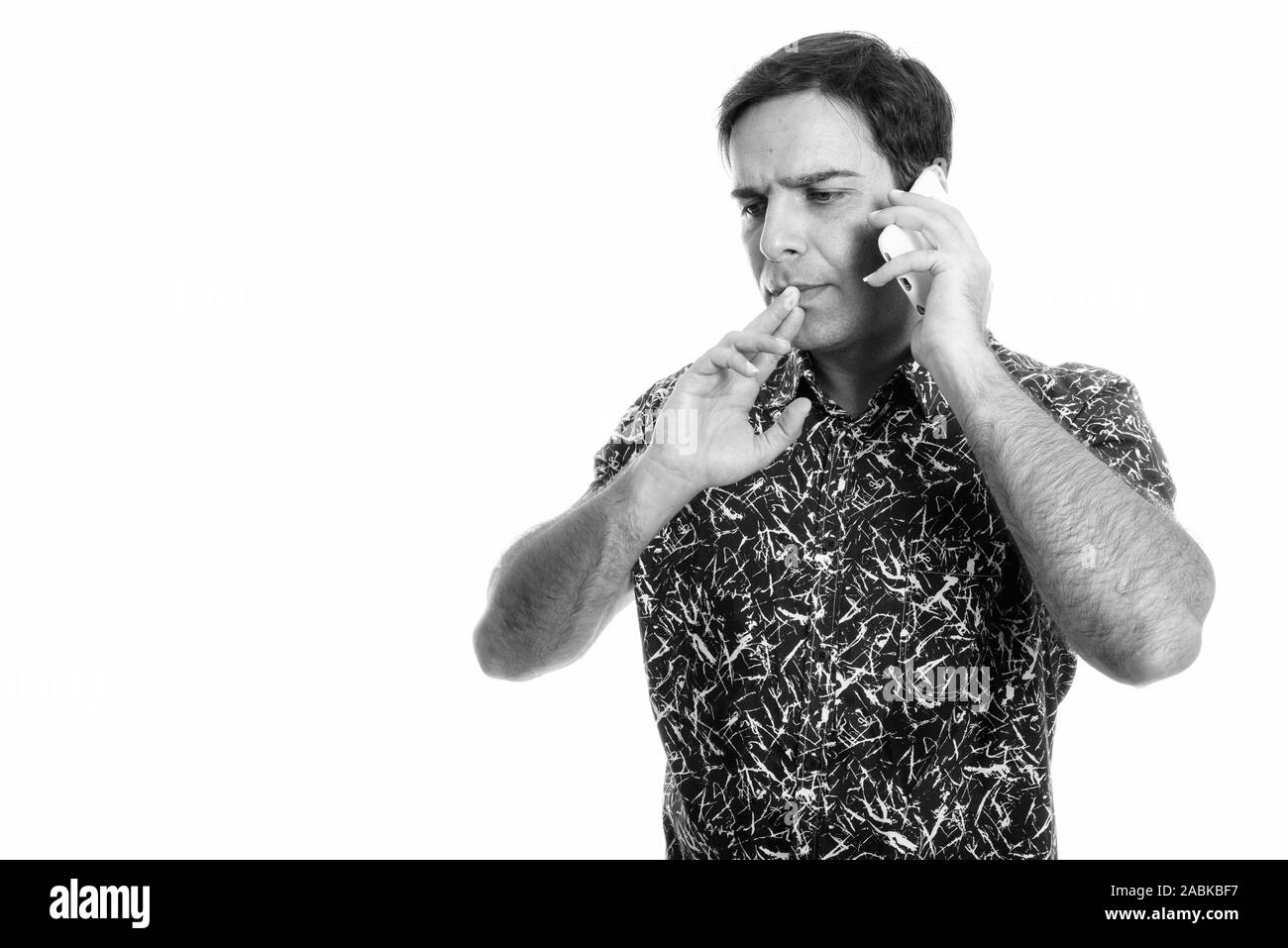 Studio shot of young Persian man thinking while talking on mobile phone isolated against white background Stock Photo