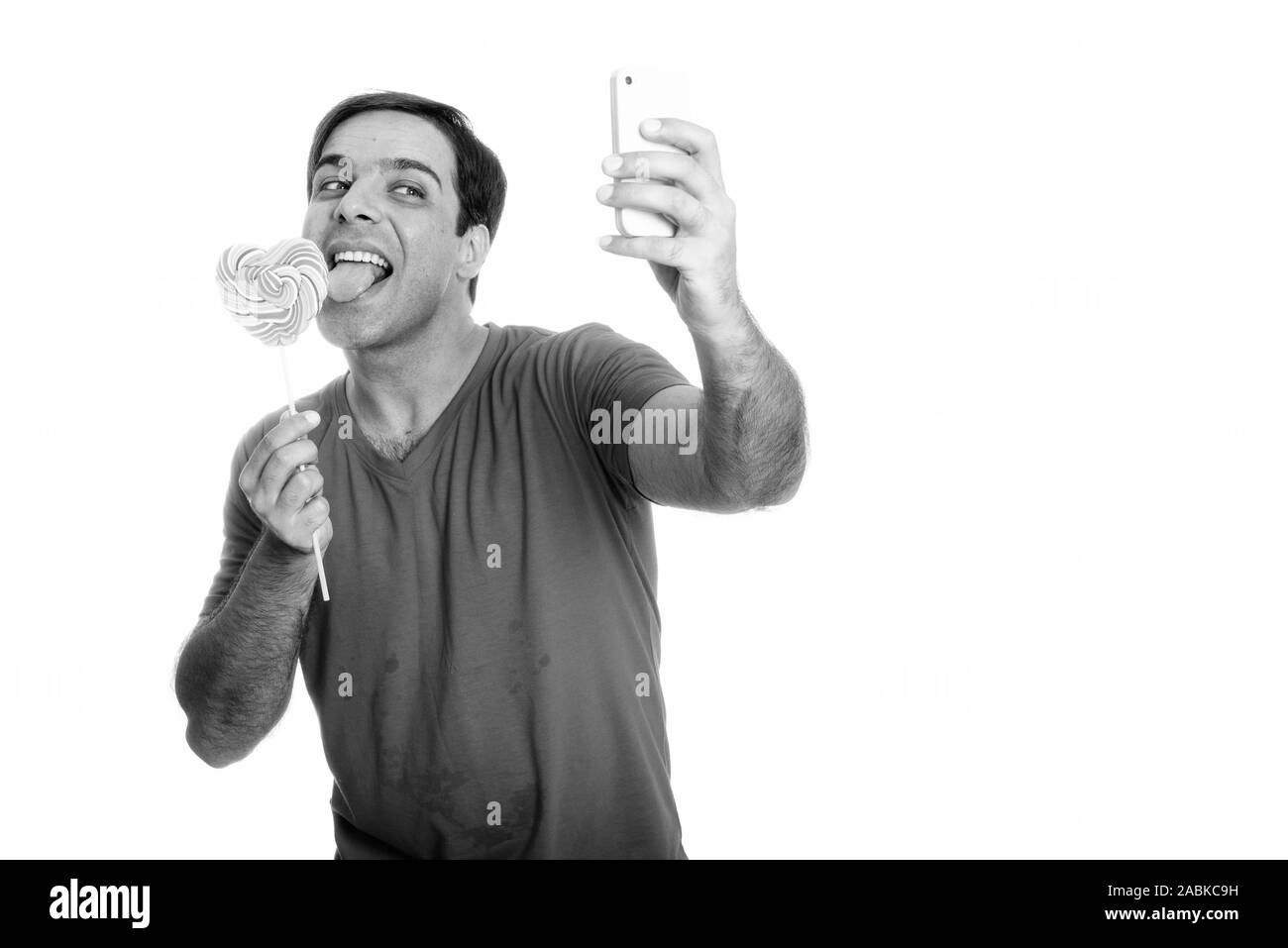 Happy Persian man smiling and taking selfie picture with mobile phone while licking heart shaped lollipop Stock Photo