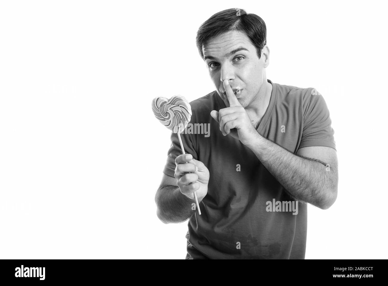 Studio shot of young Persian man holding heart shaped lollipop with finger on lips isolated against white background Stock Photo