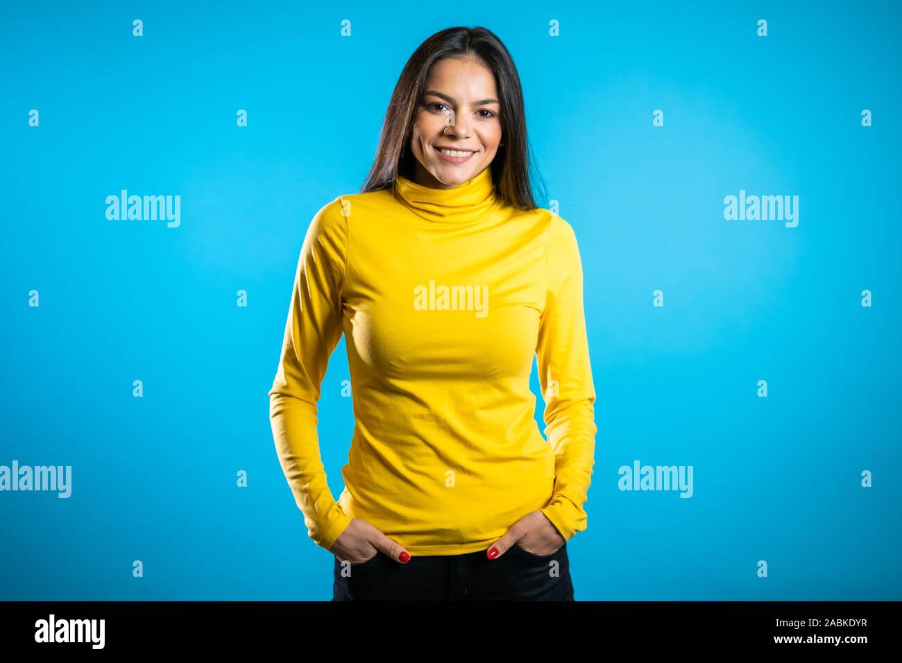Beautiful cheerful mixed race woman in yellow clothing smiling over blue wall background. Cute hispanic girl's portrait. Stock Photo