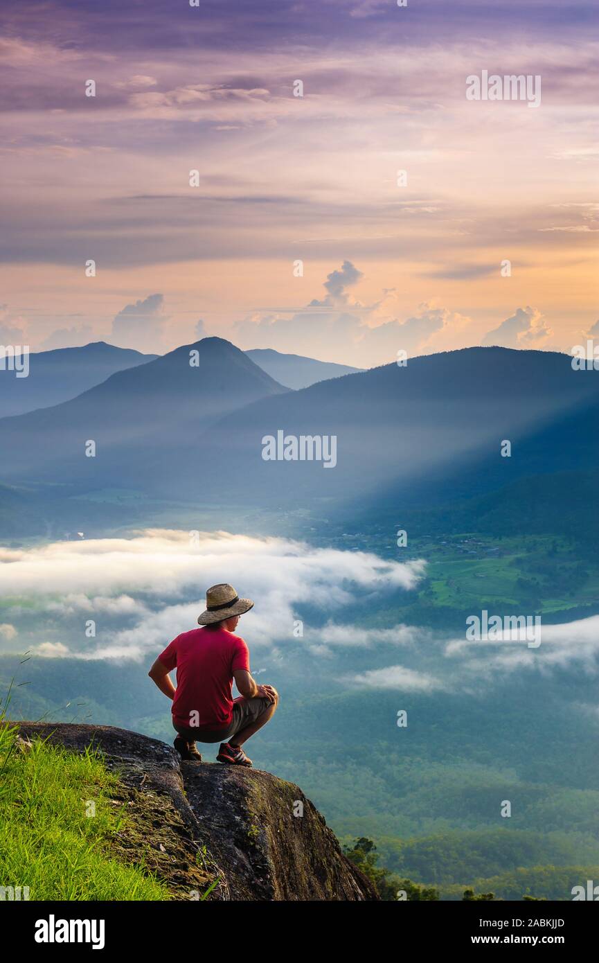 Adult Male crouching on a boulder overlooking a beautiful valley at Sunrise. Stock Photo