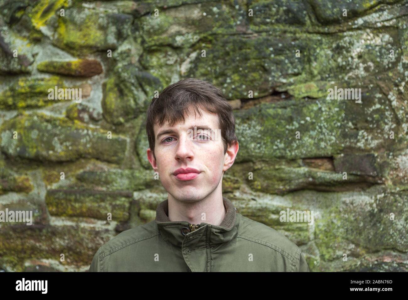 Head and shoulders of a confident and friendly young man, late teens or 20 something, standing next to a stone wall with copy space. Stock Photo