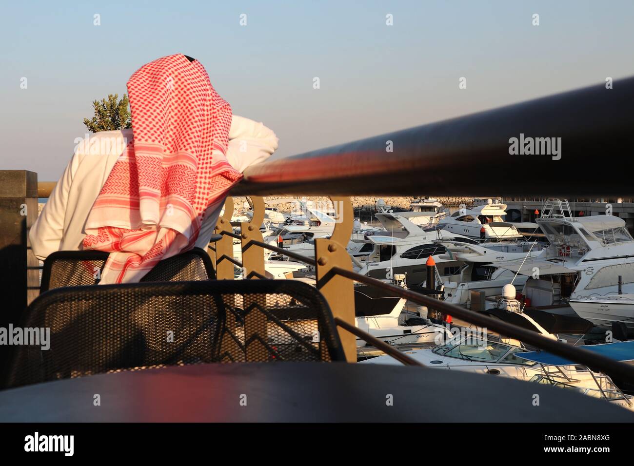 Kuwaiti man wearing red and white head dress (kaffiyeh) seen from behind sitting overlooking a marina with boats Stock Photo