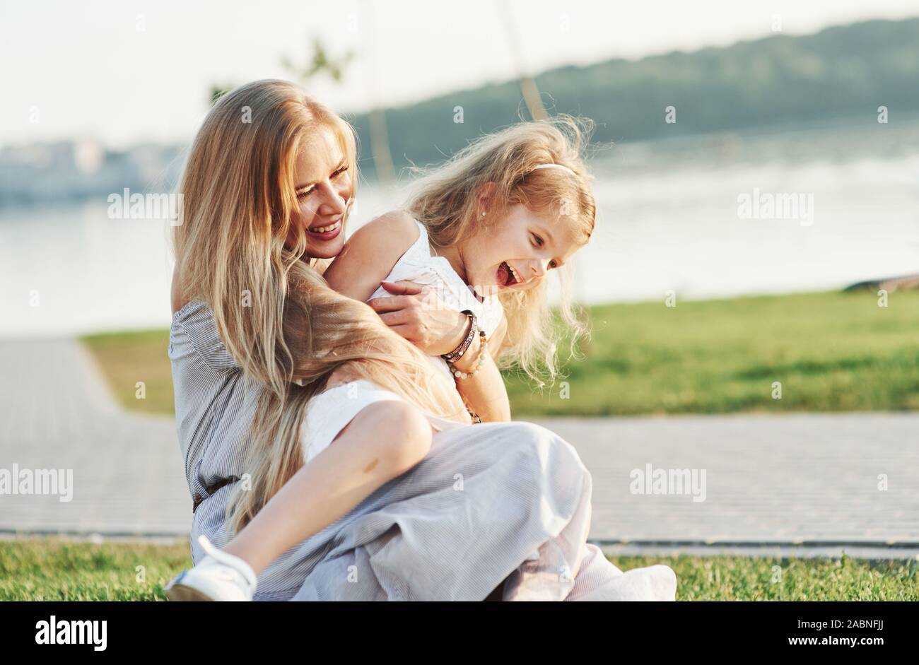 Pure love. Photo of young mother and her daughter having good time on the green grass with lake at background Stock Photo