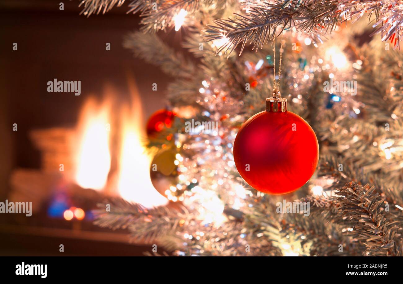 Bright red Christmas ornament hanging on silver Xmas Tree with fire place in background Stock Photo