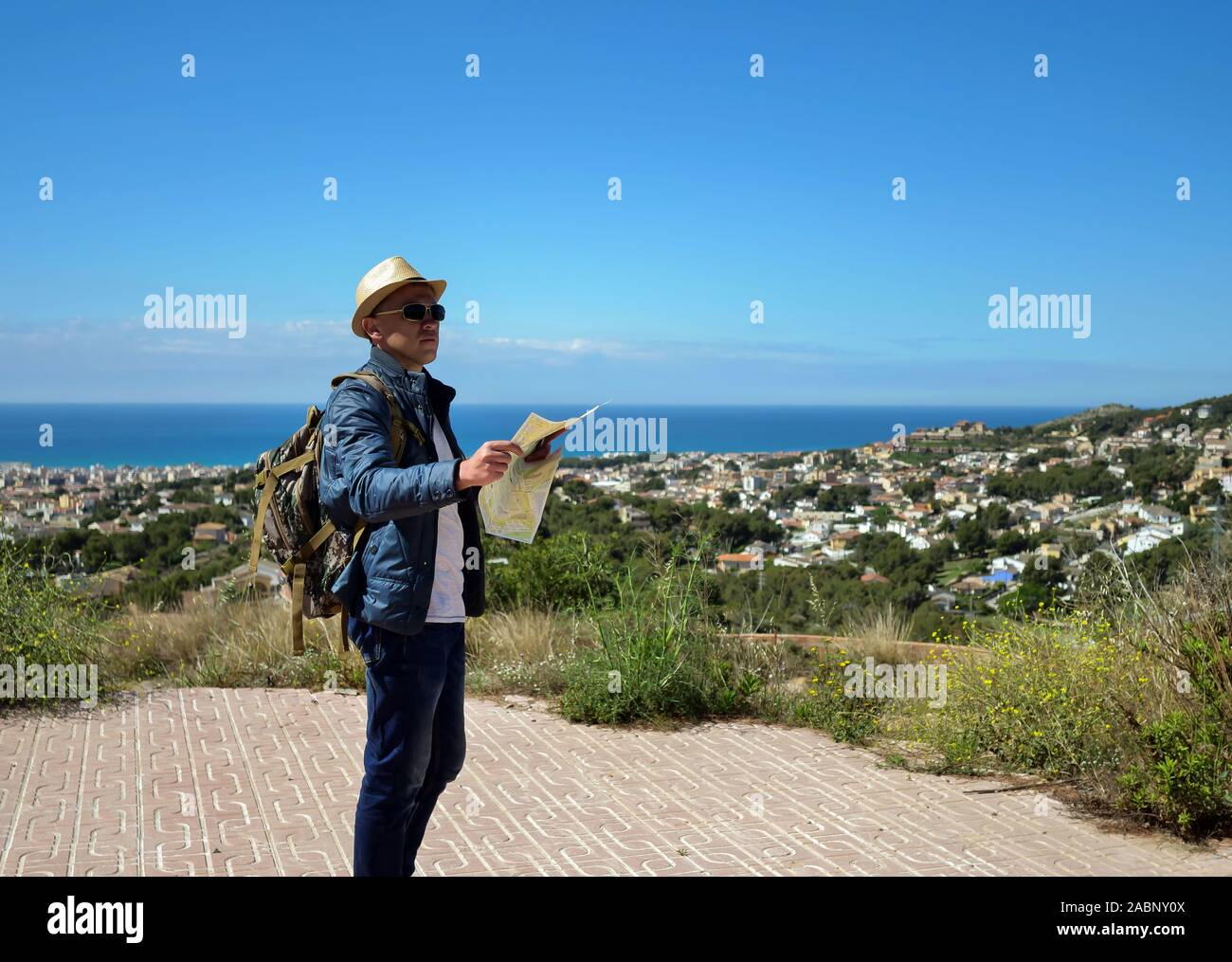 A tourist with a backpack and a map of the area in his hands stands on a mountain near the city and the sea Stock Photo