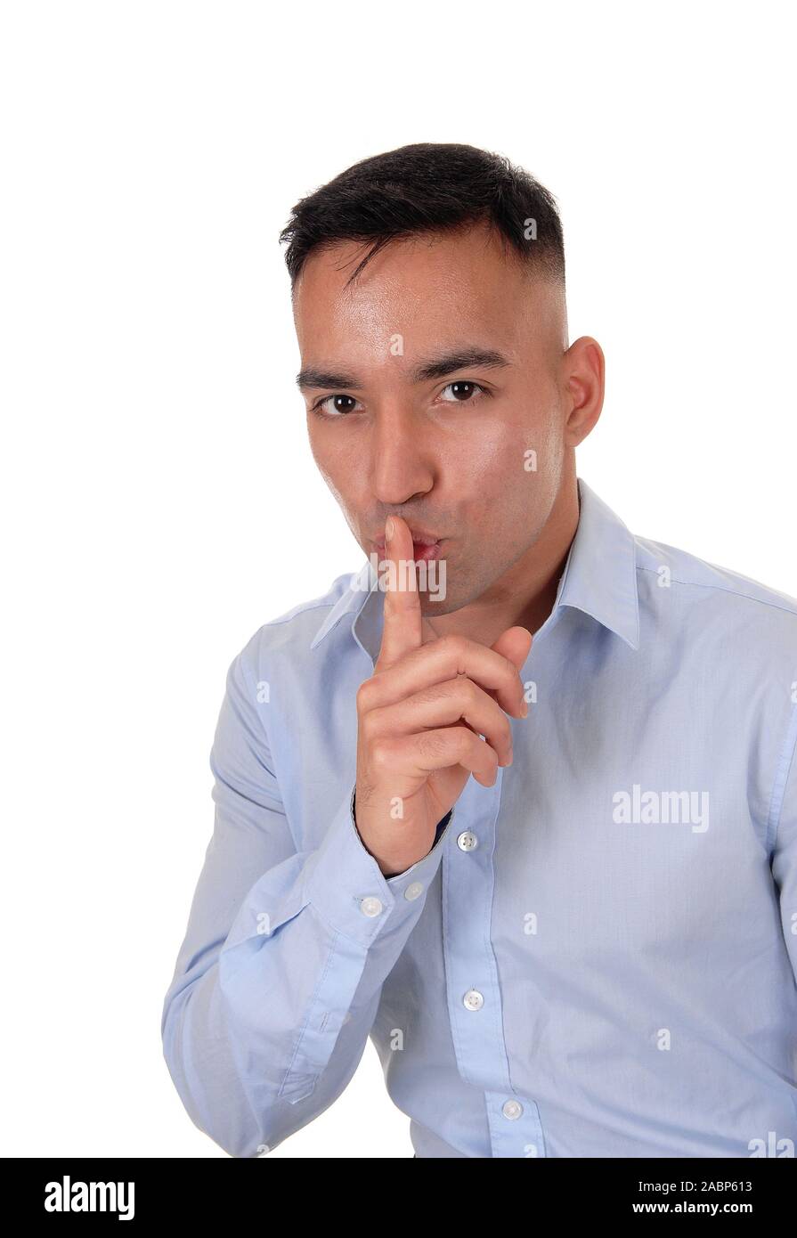 A close up image of a young East Indian man in a blue shirt with his  finger over his mouths, don't talk, isolated for white background Stock Photo