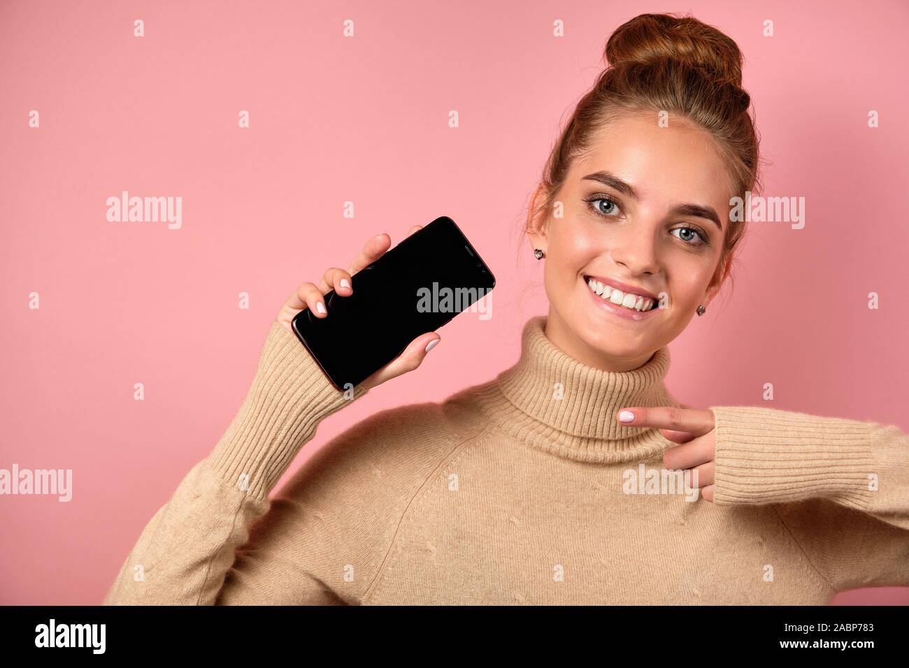 A girl with clean skin and a high bun joyfully points to the smartphone with her finger and looks at the frame. Stock Photo