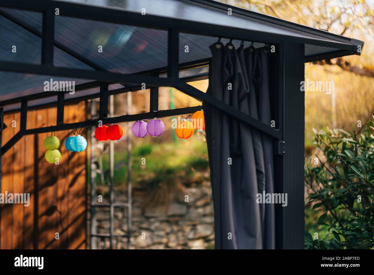 Little colorful lampion lights hanging on a pavilion in a garden on a sunny summer day. Cozy Chairs and Table standing underneath it. Stock Photo