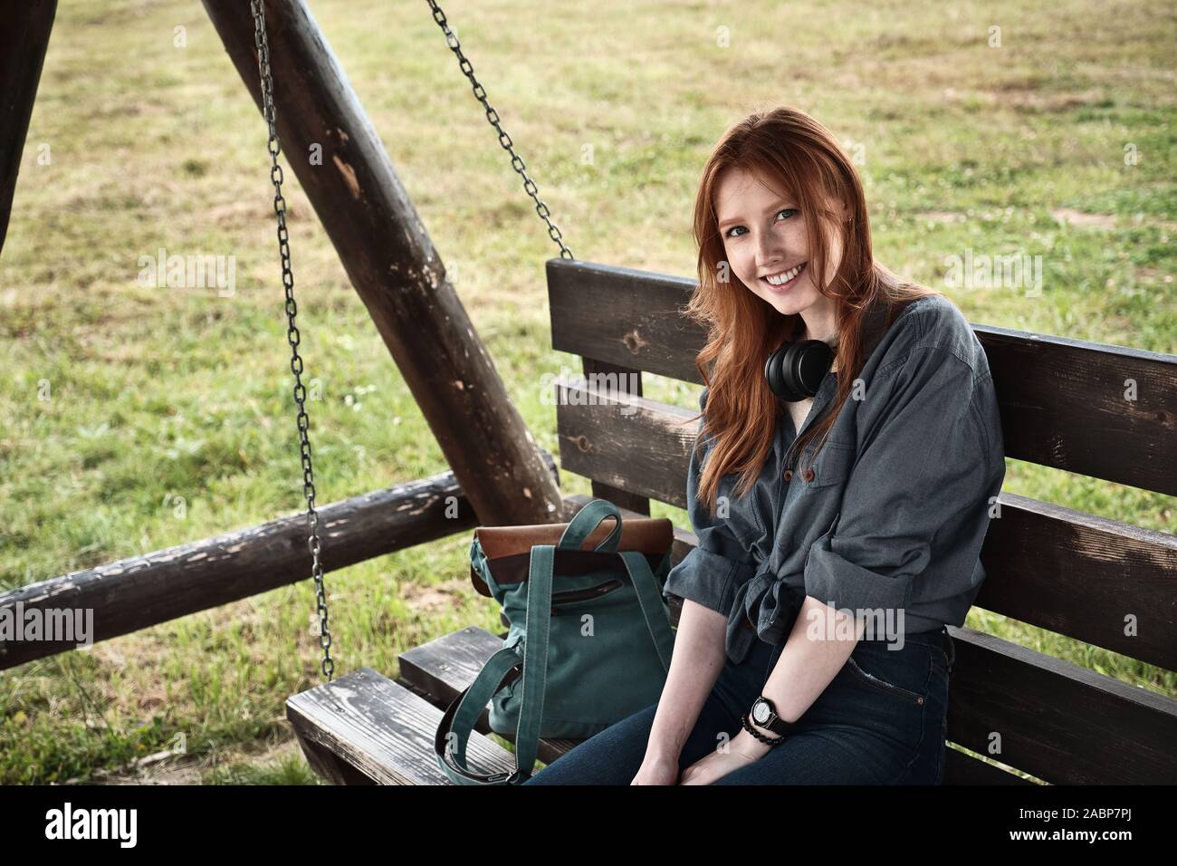 Redhead girl in a denim shirt sits with a backpack on a wooden swing bench and smiles into the frame. Stock Photo