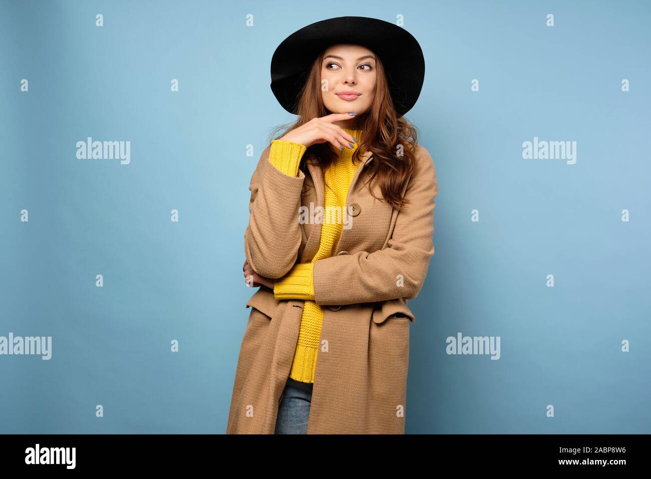 The brunette stands on a blue background in a yellow sweater, trench coat and black hat, looks away, raising an eyebrow. Stock Photo