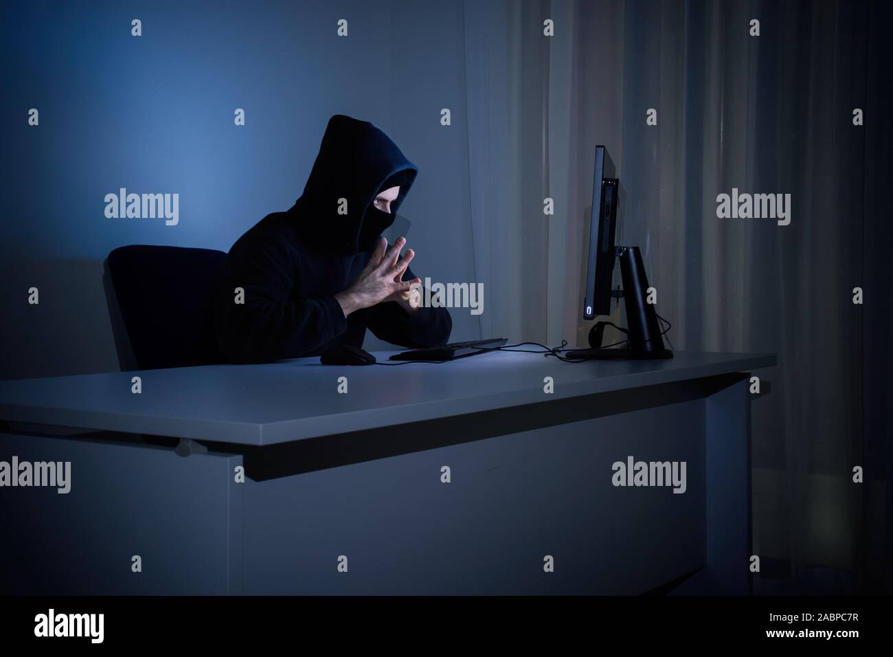 Masked hacker in front at computer into dark room represents the danger of surfing the internet. Internet security concept. Stock Photo