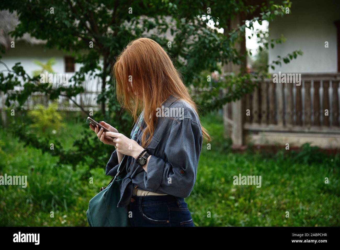 A red-haired girl in a denim shirt stands in profile with head bowed and looks at the phone against the backdrop of a green yard Stock Photo