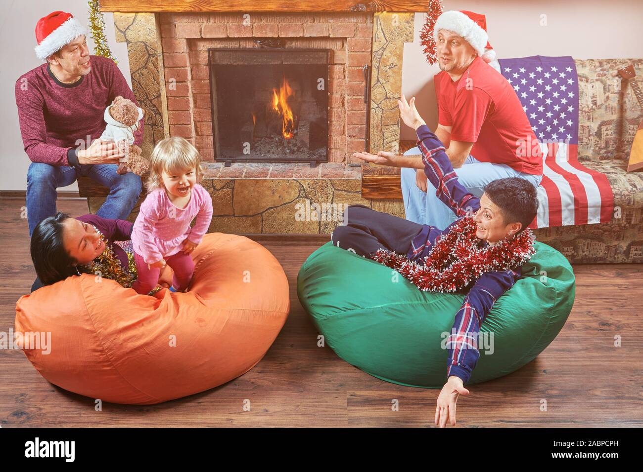 Group of people are celebrating Christmas at home in Santa's hats. Two couples of parents and their children are playing and smiling near fireplace. Stock Photo