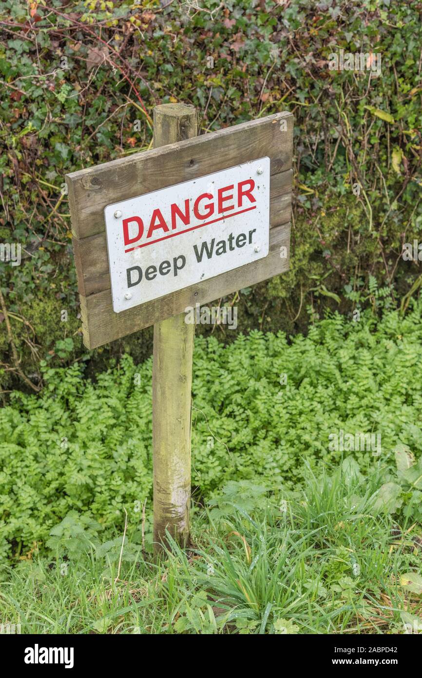 Danger warning sign for deep water drainage ditch. Plant mass may be Lesser Water-Parsnip / Berula erecta or Fool's Watercress / Apium nodiflorum. Stock Photo