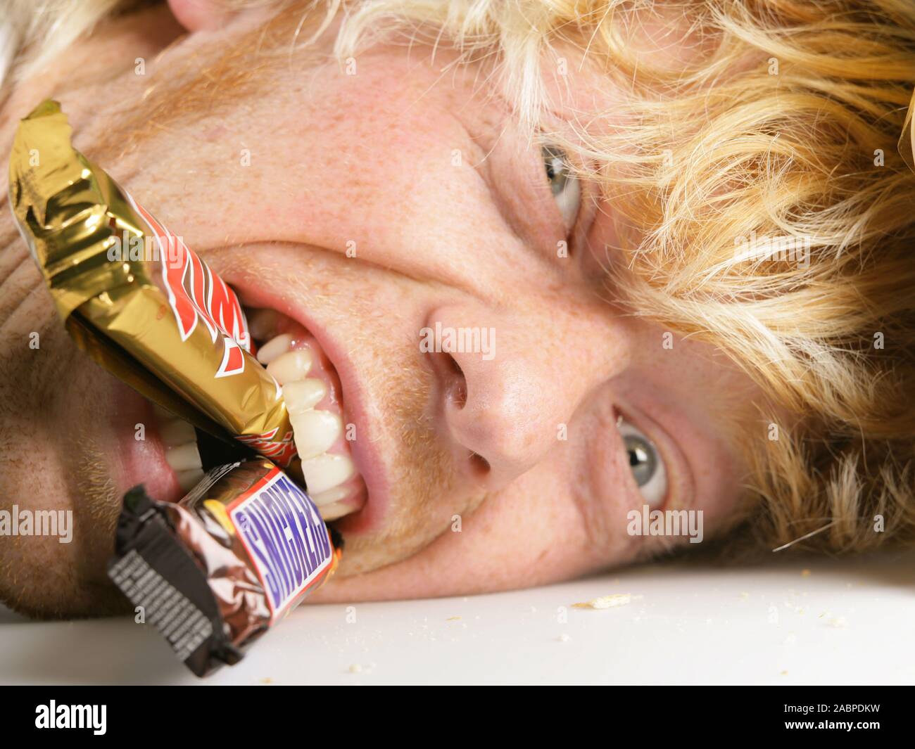 Blonder junger Mann beisst mit Genuss in Schokoladenartikel - Vernarrt in Suessigkeiten - Symbolfoto - Suchtgefahr Junger Mann schneidet Grimassen Stock Photo