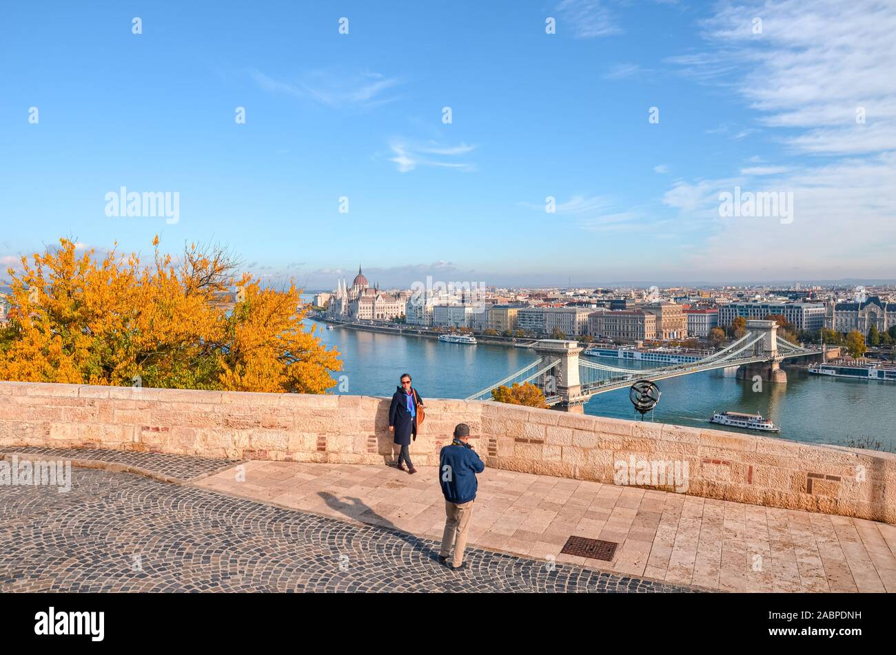 Budapest, Hungary - Nov 6, 2019: Older Asian couple taking travel pictures on the viewpoint above the Hungarian city. Danube river, Hungarian parliament building and Szechenyi Bridge in background. Stock Photo