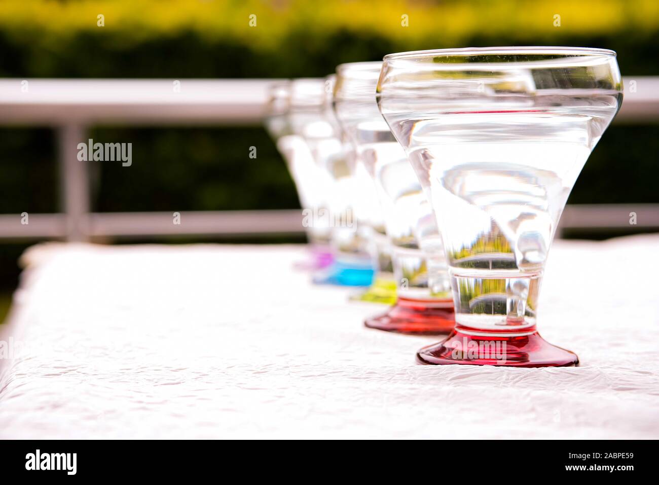 Colorful drinking glasses on white background standing in a row in natural light with green foliage background. red, orange, blue green purple . Stock Photo