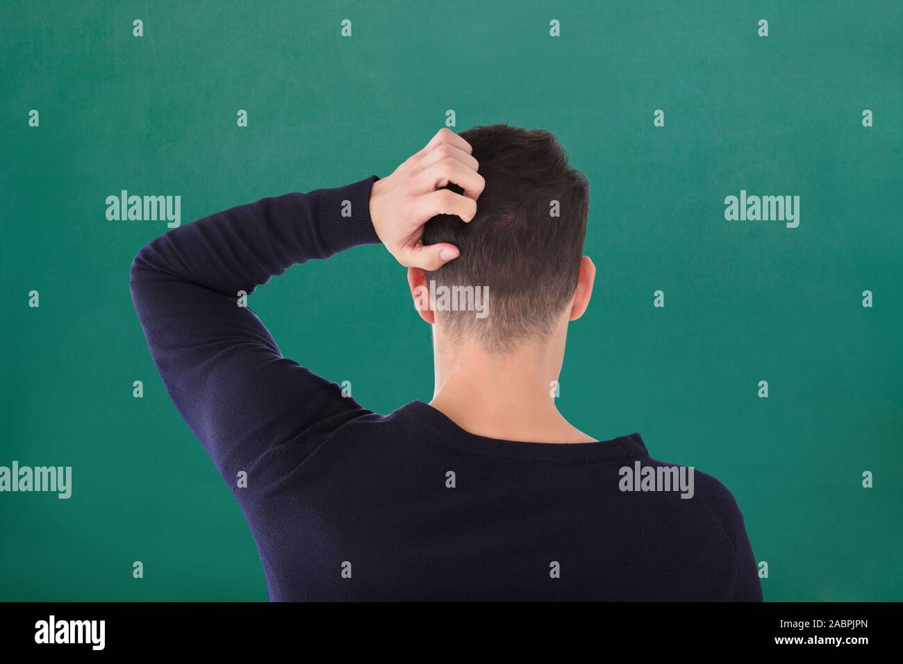 Rear View Of Young Man In Blue T-shirt Scratching His Head Against Green Background Stock Photo