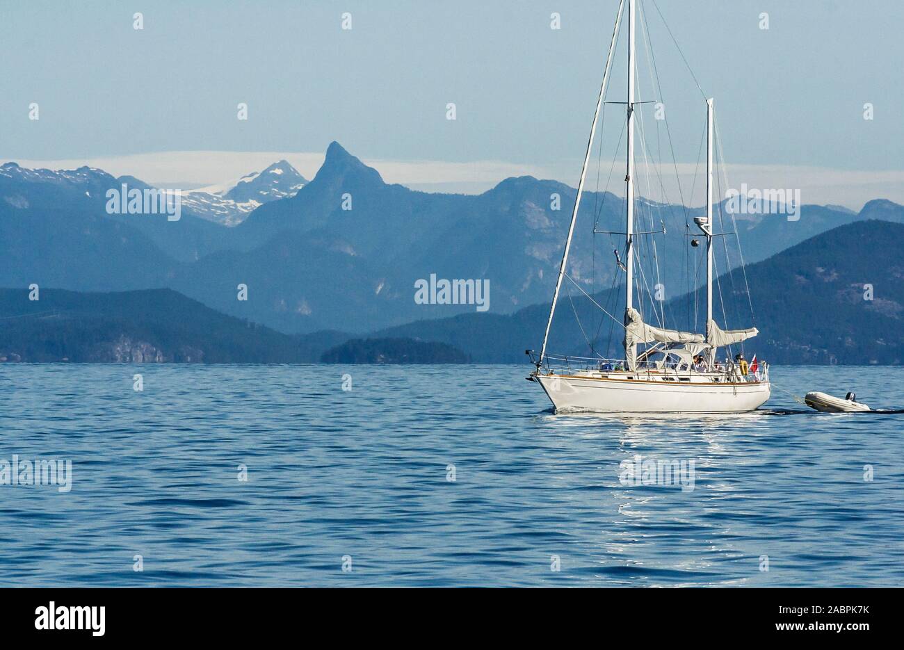 A Canadian sailboat is underway, motoring along on a beautiful summer's day in BC's Desolation Sound, with steep mountains rising in the background. Stock Photo