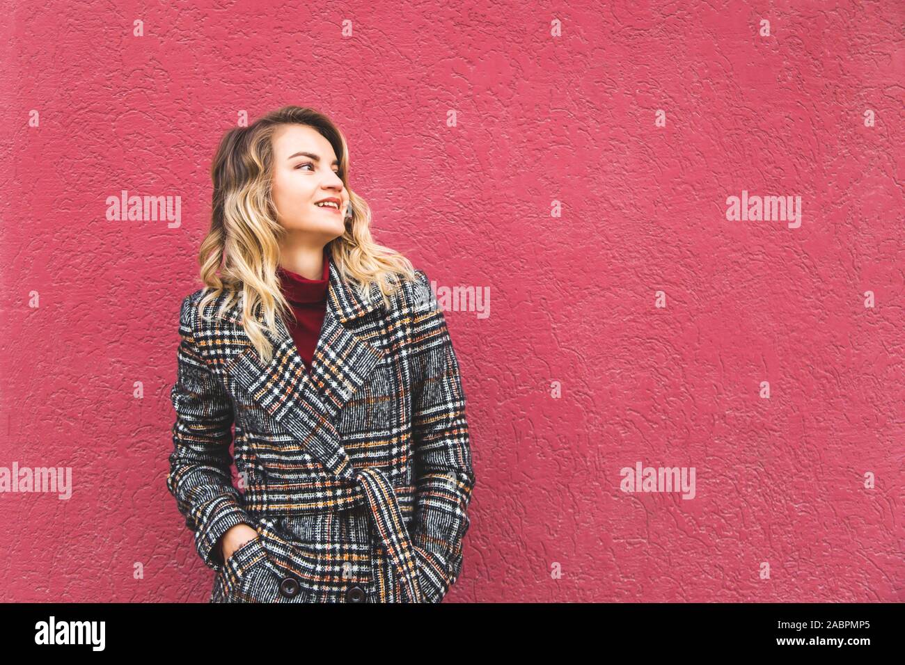 A modern stylish Caucasian girl in a plaid coat against the background of a bright wall looks thoughtfully to the side. Stock Photo