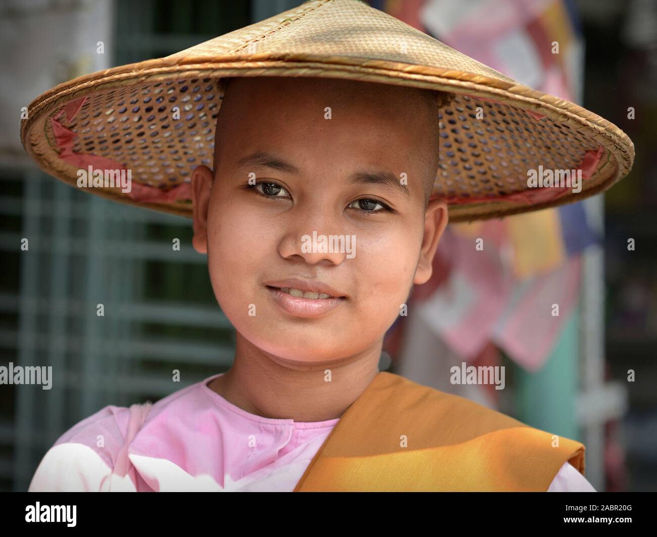Young Burmese Buddhist nun wears an Asian conical straw hat and smiles for the camera. Stock Photo