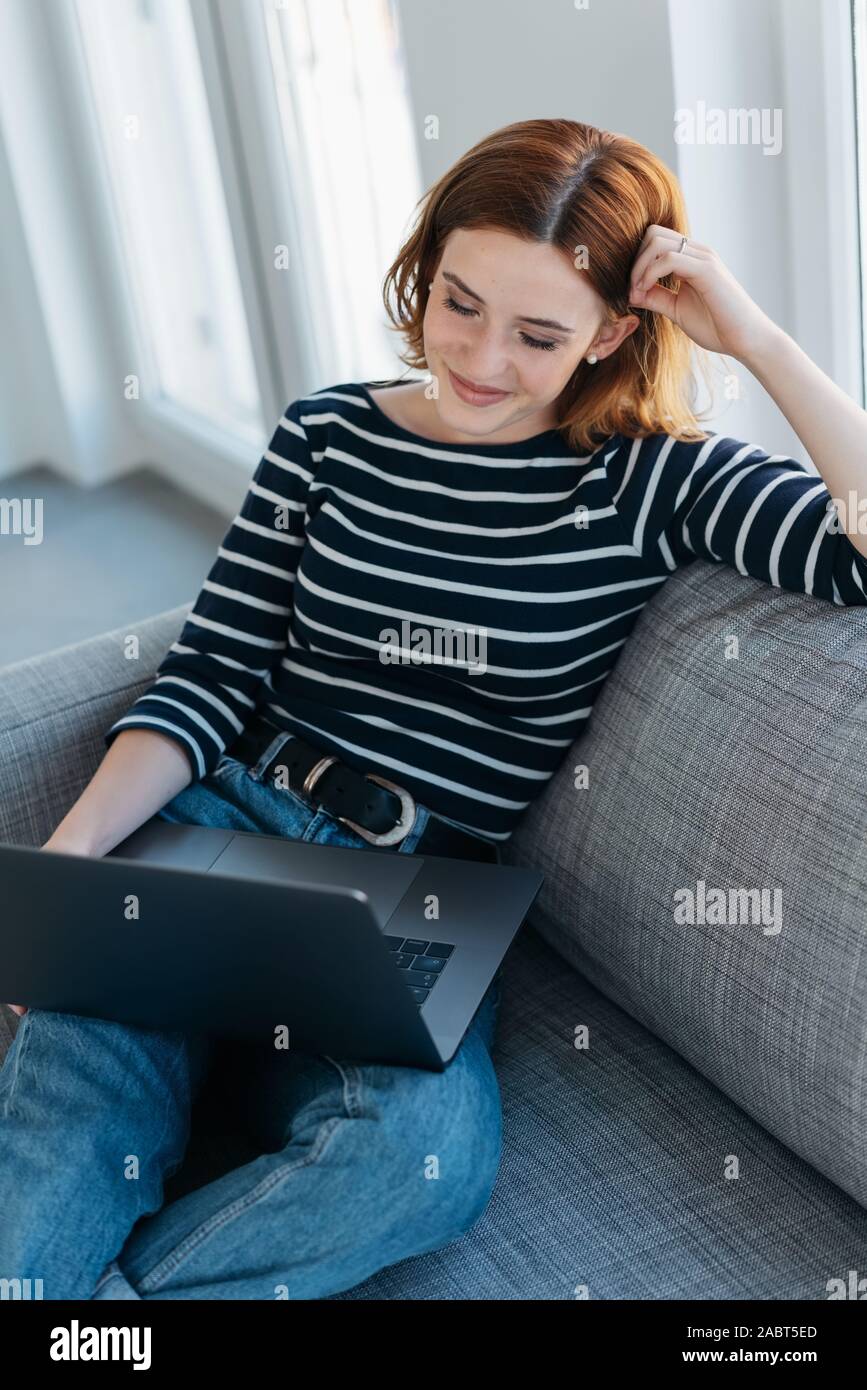 Contented young woman unwinding at home relaxing on a comfortable sofa with her laptop Stock Photo