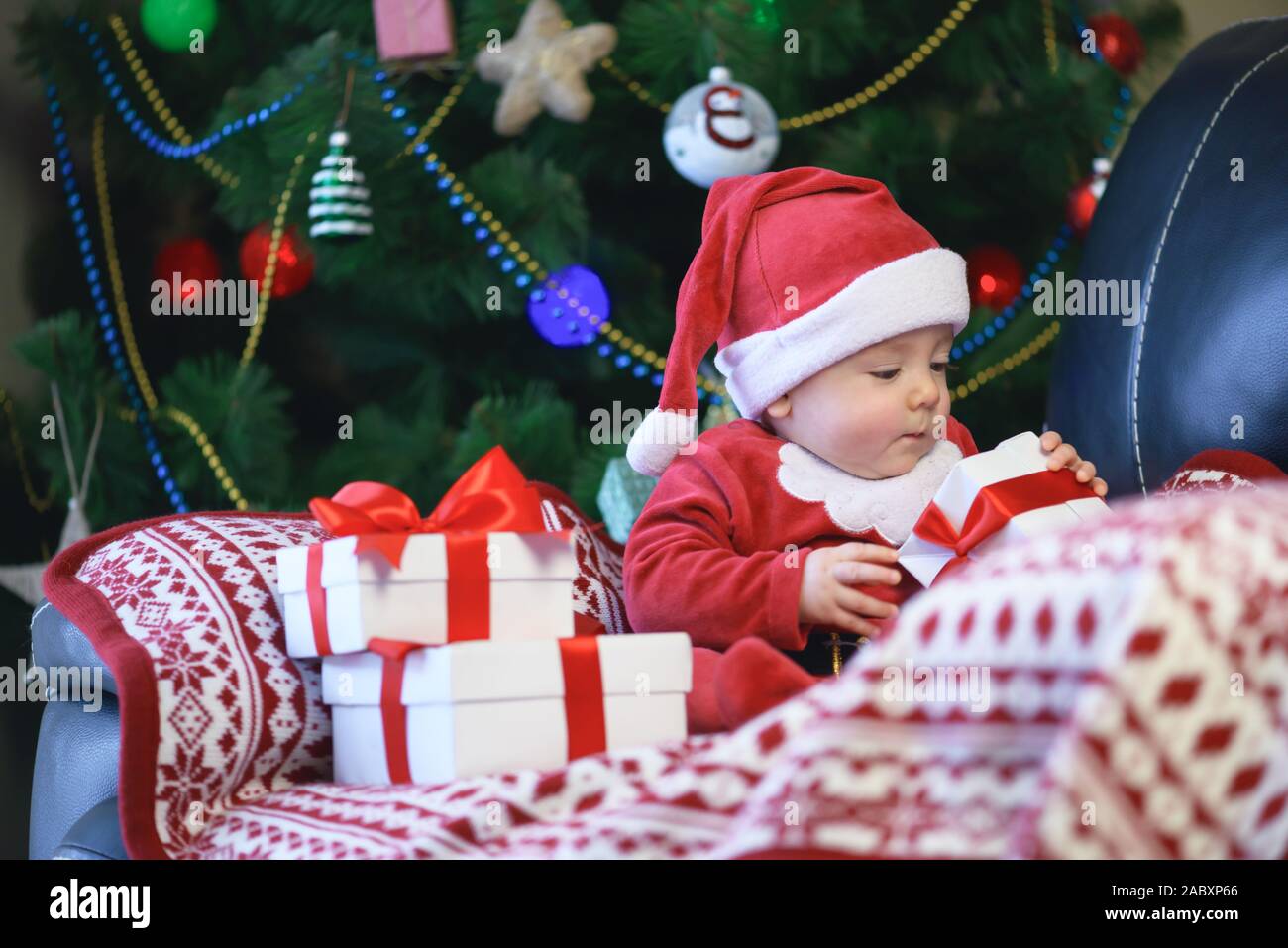 Baby boy in Santas clothes holding Christmas present gift box in front of Christmas tree Stock Photo