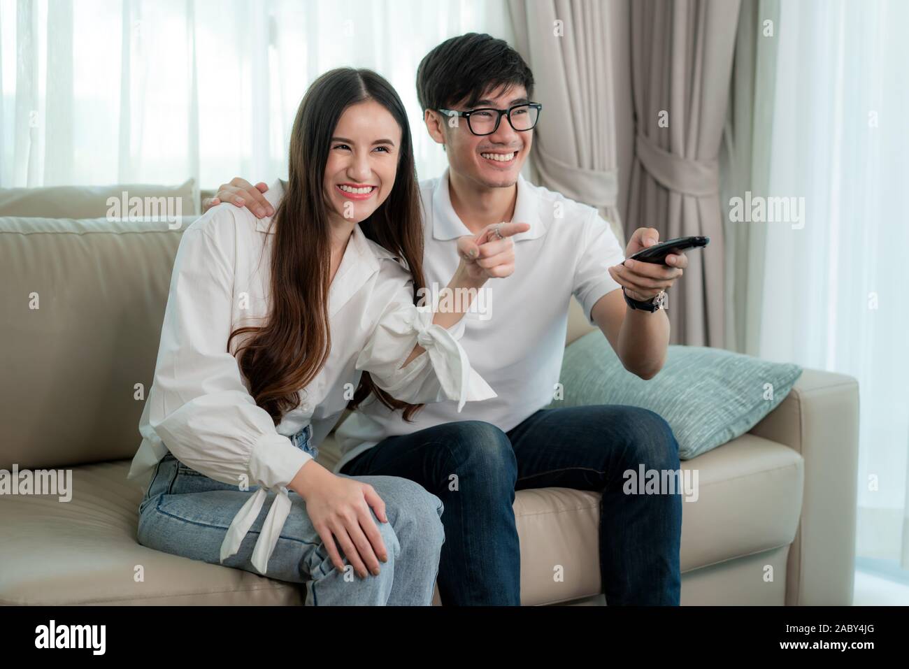 Asian young loving couple sitting and watching TV and smile when watching fun program in TV in a comfortable couch in the livingroom at home. Family l Stock Photo