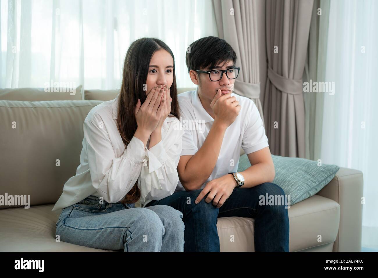 Asian Couple man and woman watching and enjoying terror tv movie sitting on a couch together in the livingroom at home. Family lifestyle relax and rec Stock Photo