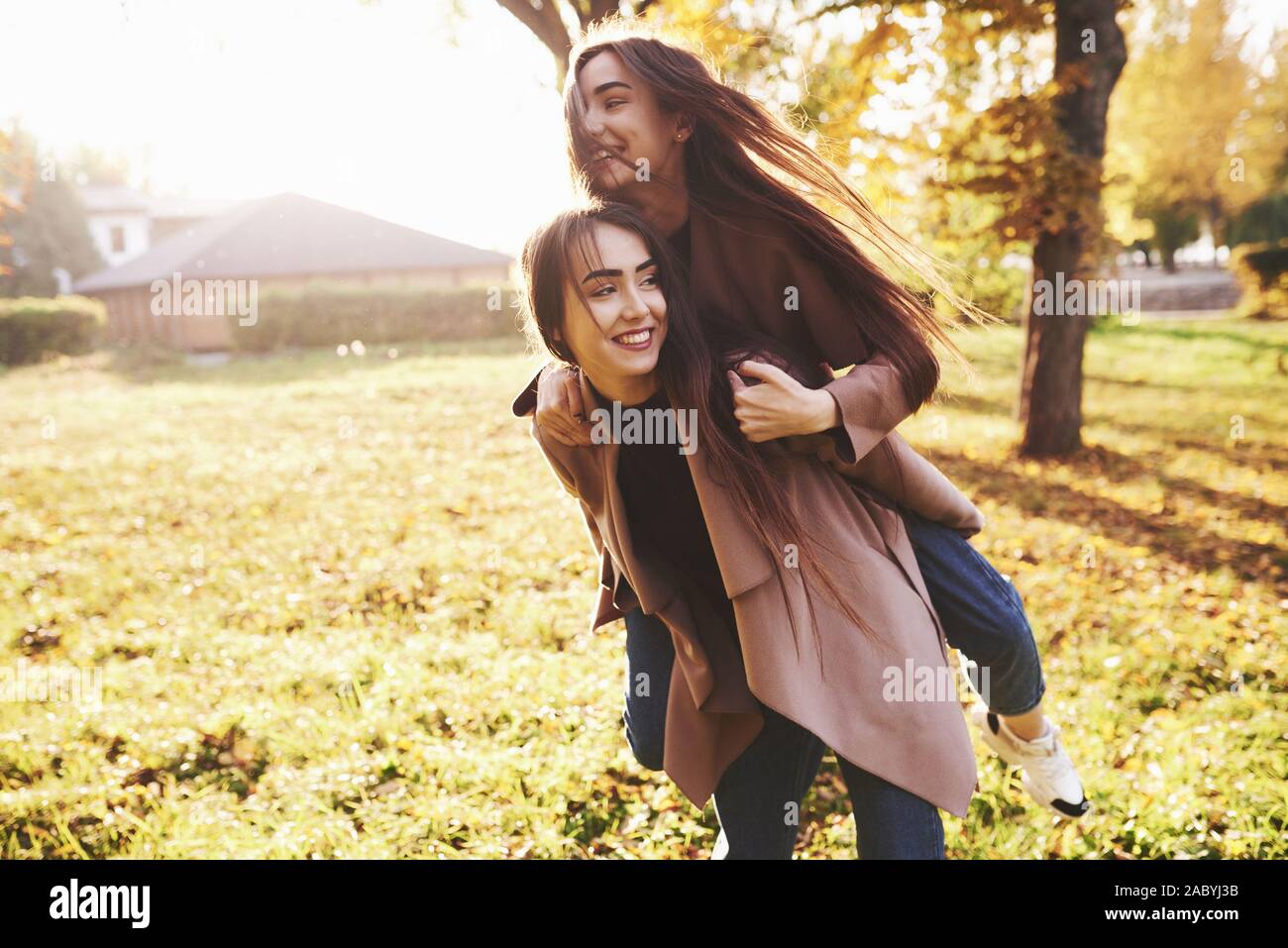 Young smiling brunette twin sisters having fun and doing a piggy back riding in autumn sunny park on blurry background Stock Photo